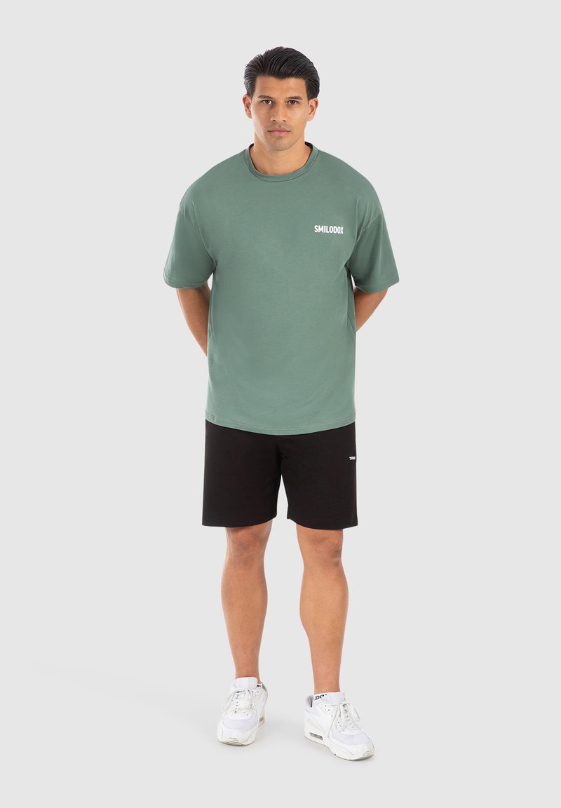 Grøn bomulds T-shirt med korte ærmer og hvidt logo på brystet, parret med sorte shorts og hvide sportssko.