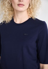Tamsiai mėlynas medvilninis marškinėliai su apvalia iškirpte, trumpomis rankovėmis ir tekstūruota juoda logotipu kairiajame krūtinės šone. Lygus, minimalistinis dizainas.