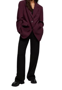 Vinröd oversized blazer med strukturerade axlar, enkel knappstängning och slag med hack, kombinerad med vida svarta byxor och klumpiga skor.