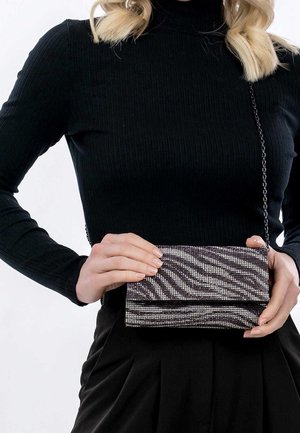 Kobieta w czarnym prążkowanym golfie i plisowanej spódnicy trzymająca małą fioletowo-srebrną torebkę w paski zebry z łańcuszkowym paskiem.