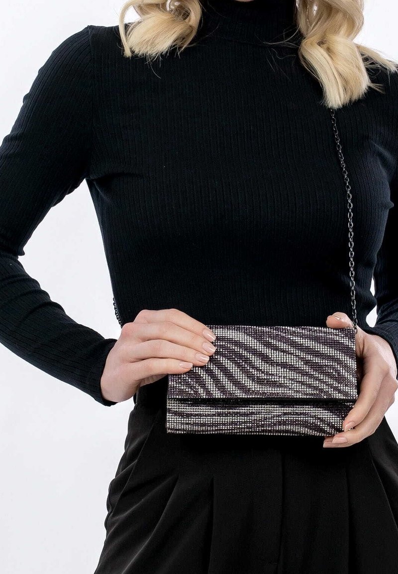 Femme portant un col roulé noir côtelé et une jupe plissée, tenant une petite pochette violette et argentée à rayures zébrées avec une bandoulière chaîne.