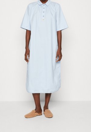 Robe bleu clair, longueur genou, avec des manches courtes, un col et des boutons. Portée avec des chaussures mules en peluche de couleur tan.