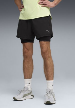 Homme portant un short superposé noir Puma, des chaussettes blanches Puma et des chaussures de course blanches Puma, debout les mains sur les hanches sur un fond gris.