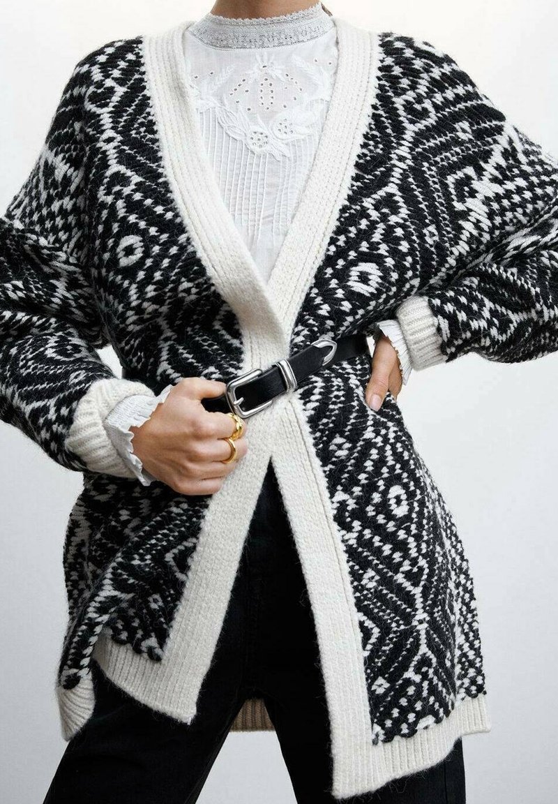 Personne portant un cardigan en maille à motifs noir et blanc sur un chemisier blanc brodé, ceinturé à la taille avec une ceinture noire et des bagues en or à la main.