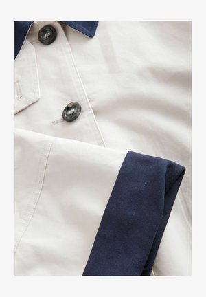 Bežinė medžiaga su tamsiai mėlynomis apykaklės ir rankovių akcentais; su juodais mygtukais ir subtiliomis siūlėmis, sukurianti švarų, struktūrizuotą įvaizdį.