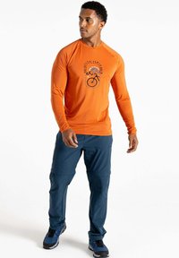 Langärmliges oranges Hemd mit einem Fahrrad-Graphic, kombiniert mit blauen Cargo-Hosen. Das Hemd hat einen enganliegenden Schnitt und eine glatte Textur.