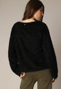 Vrouw met lang bruin haar, gekleed in een zachte zwarte trui en olijfgroene broek, met een lichte draaiing weg van een neutrale achtergrond.