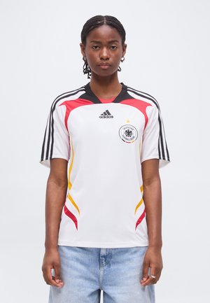 Jeune femme aux cheveux tressés portant un maillot de football blanc Adidas Allemagne avec des accents rouges et jaunes et un jean bleu clair.