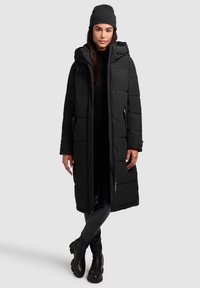 Black puffer coat with a hood, featuring a quilted design, front buttons, and side pockets. Model wears a matching black beanie and boots.