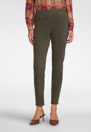Femme portant un pantalon slim vert olive, des chaussures à talons marron avec bride arrière et une chemise à manches longues à carreaux beige et rouge, debout sur un fond blanc.