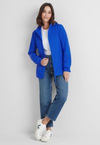 Bright blue lightweight rain jacket with a hood, elastic cuffs, front zip closure, and drawstring at the hem, worn over a white shirt and jeans.