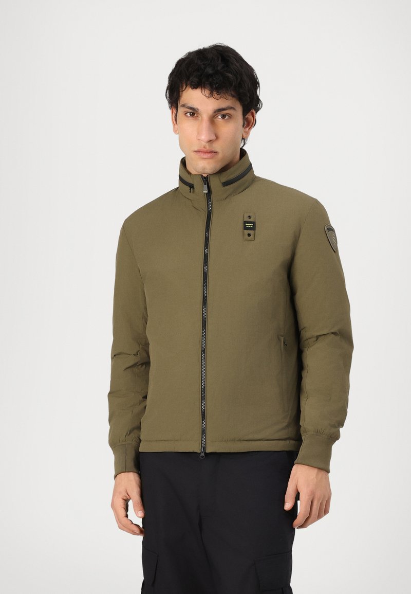 Veste vert olive avec un col haut, fermeture éclair à l'avant et accents noirs. Présente des poignets élastiques et un patch logo sur l'épaule.