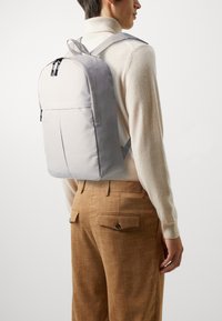 Lys grå ryggsekk med avrundet topp, glatt stoff, doble glidelåser og justerbare stropper, båret over en beige genser og brune bukser.