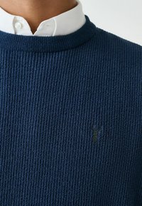 Nahaufnahme einer Person, die ein weißes Hemd mit Kragen unter einem strukturierten, dunkelblauen Pullover trägt, der ein kleines gesticktes Logo auf der Brust hat.