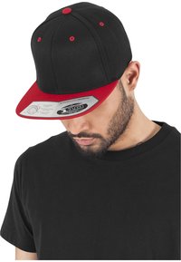 Sort baseball cap med rød skygge og detaljer. Har en struktureret form og justerbar snapback-lukning. Etiket viser produktspecifikationer.