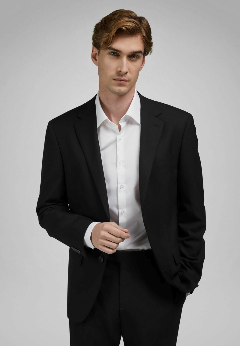 Jonge man met lichtbruin haar, gekleed in een zwart pak en wit overhemd, poseert met één hand gedeeltelijk in de zak tegen een grijze achtergrond.