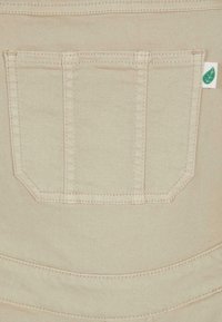 Beige denimstoff med en baklomme med vinklet hjørner, overlockede detaljer og et lite grønt bladmerke.