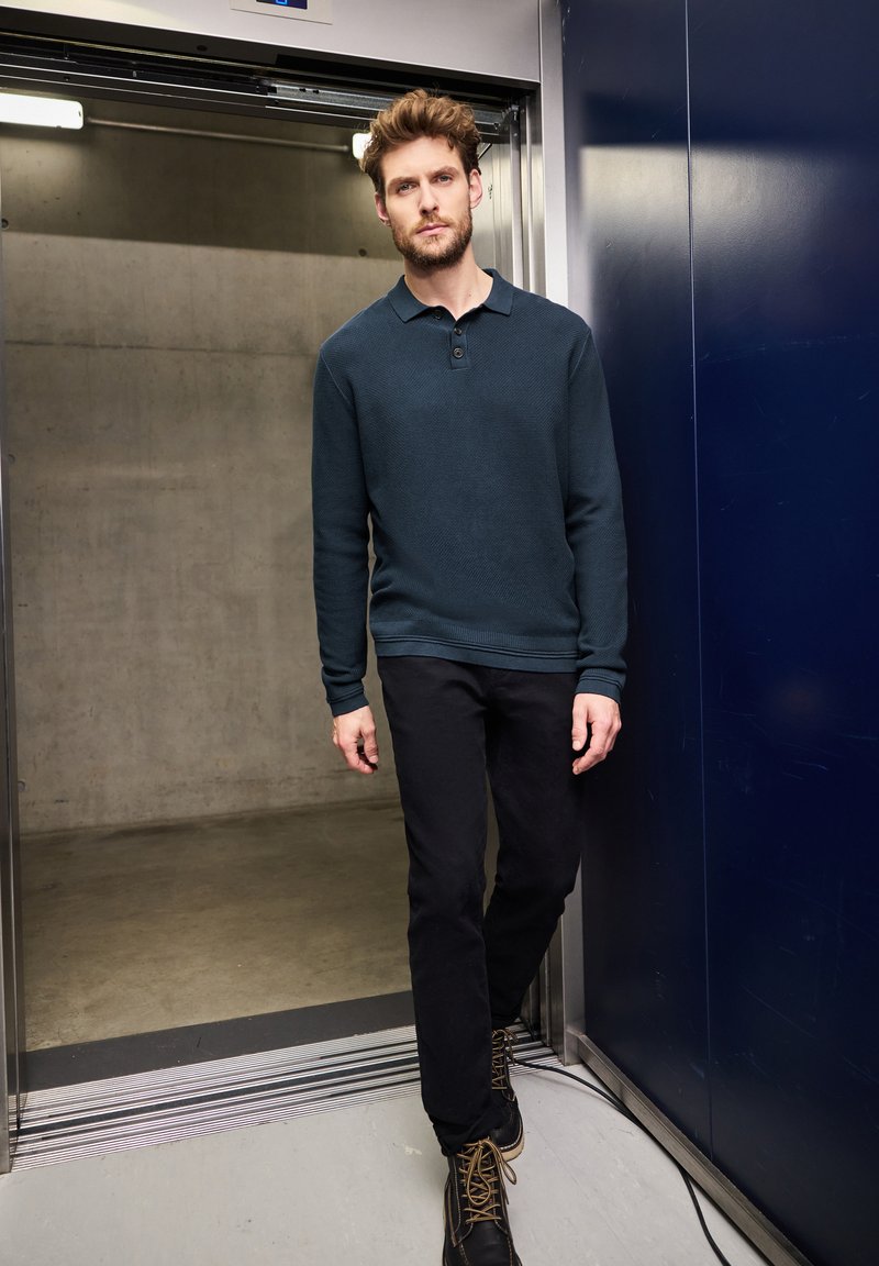 Dunkelblauses Langarm-Polohemd mit strukturiertem Muster, zwei-Knopf-Blende, kombiniert mit schwarzen Slim-Fit-Hosen und braunen Stiefeletten.