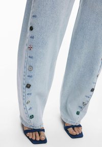 Helle blaue Jeans-Weitbein-Hosen mit dekorativen Knöpfen entlang der Seitennaht. Der Stoff hat eine glatte Textur mit sichtbaren Nähten.