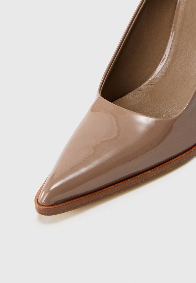 Zapato de tacón alto de charol beige con punta afilada y corte lateral bajo, que presenta una textura suave y una suela de goma color marrón.