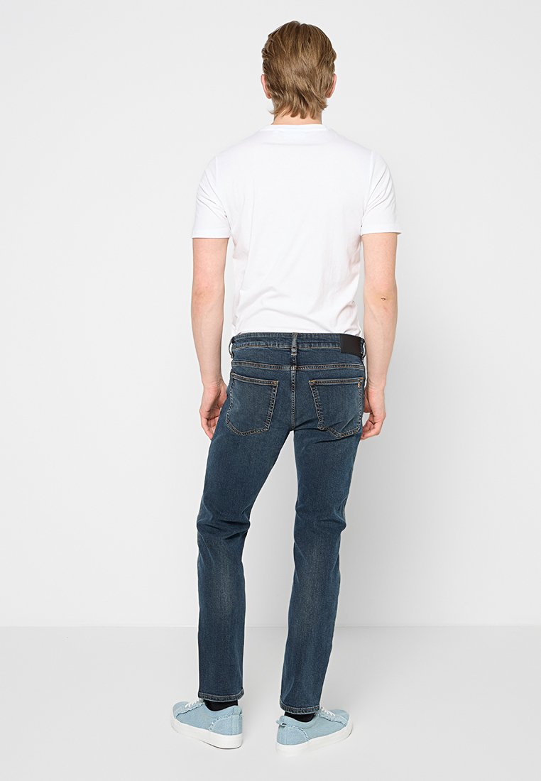 Jeans en denim bleu foncé avec une coupe droite, comportant deux poches arrière et un petit patch logo à la taille. Portés avec un polo blanc.