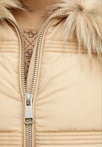 Beige polstret jakke med en fremtrædende sølv lynlås og faux pels kanter. Piping-detalje forbedrer designet. Indersiden har et tekstureret mønster.