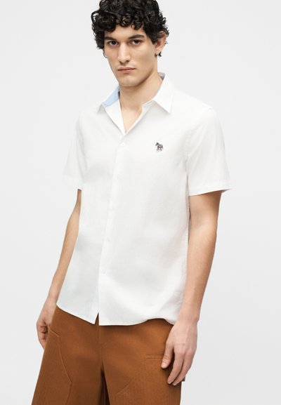 Young man with curly hair wearing a white short-sleeve button-up shirt with a small embroidered zebra emblem and brown pants.