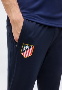 Main dans la poche, portant un pantalon de sport bleu marine avec le blason du club Atlético Madrid sur la cuisse.