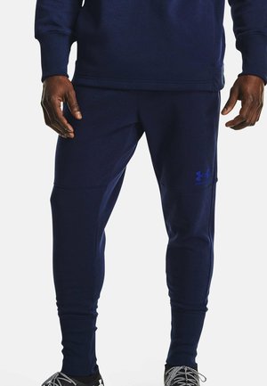 Homme portant un jogging Under Armour bleu foncé et un haut assorti, debout avec les mains détendues le long du corps devant un fond uni.