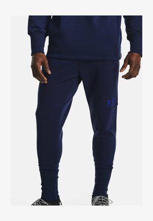 Homme portant un jogging Under Armour bleu foncé et un haut assorti, debout avec les mains détendues le long du corps devant un fond uni.