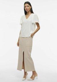 Blusa bianca con maniche a sbuffo, abbinata a una gonna beige di lunghezza media con uno spacco frontale e tasche. Mules con tacco trasparente completano il look.