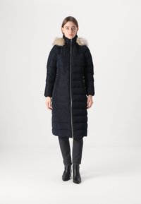 Lungo piumino nero con zip frontale e tasche laterali, dotato di cappuccio rimovibile foderato in pelliccia, con un design trapuntato.
