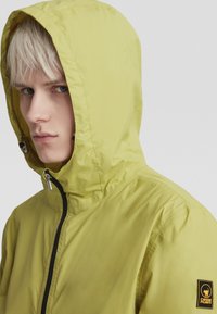 Hoodie in leuchtendem Limettengrün mit glatter Textur, ausgestattet mit einem zentralen schwarzen Reißverschluss und einem Logopatch am Ärmel.