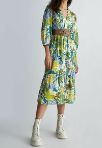 Vestido de estampado floral con escote en V, mangas abullonadas y diseño en capas. Cinturón marrón con tachuelas que acentúa la cintura. Combinado con botas blancas de cordones.