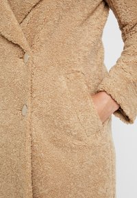Cappotto teddy beige e testurizzato con colletto a scaurtura, due bottoni e una tasca laterale a sinistra. Materiale morbido e fluffy con un design casual.