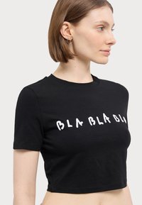 Mustan lyhythihaisen puuvillat-paidan, jossa on rohkea valkoinen "BLA BLA BLA" -printti. Pyöreä kaula-aukko. Sileä pinta.