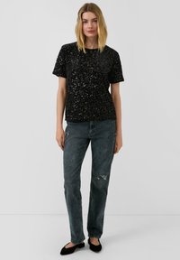 Zwarte paillettentop met korte mouwen, gecombineerd met vervaagde grijze rechte jeans met een gescheurd detail, gedragen met zwarte platte schoenen.