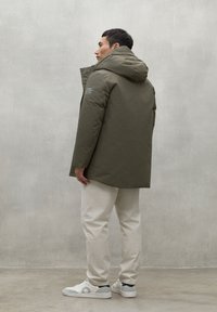 Chaqueta parka verde oliva con capucha, que cuenta con un cierre de cremallera y un diseño de marca minimalista, combinada con pantalones blancos y zapatillas blancas discretas.
