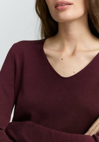 Bordeaux langarm gebreide top met een V-hals, met een geribde structuur en zichtbare stiksels langs de halslijn. Gladde stofdetails.