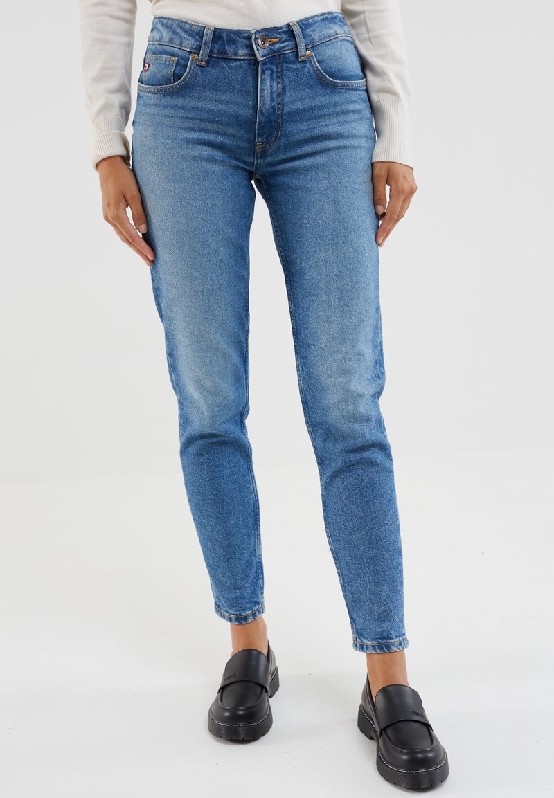 Jean droit taille haute bleu clair avec cinq poches, fermeture à boutons et texture délavée ; associé à des chaussures noires à enfiler.