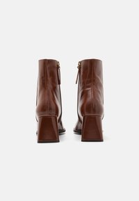 Botas de tornozelo em couro castanho com bico arredondado, salto grosso, fechos de correr laterais e acabamento liso. Detalhes ajustáveis nas alças dos fechos de correr visíveis.