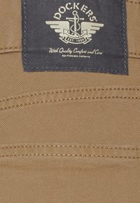 Beiger Stoff von Dockers-Hosen mit einem marineblauen Etikett, das ein Anker- und Flügellogo zeigt, gegründet 1986, mit klaren Nähdetails.