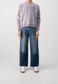 Vaalean violetti huppari, jossa on teksti "DO NOTHING CLUB", yhdistettynä leveälahkeisiin sinisiin farkkuihin, jotka näyttävät haalistuneilta. Asun viimeistelevät valkoiset ja harmaat tennarit.