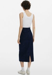 Kvinna med kort lockigt hår, iklädd en vit ärmlös topp och en mörkblå midiklänning med bakfickor och slits bak. Hon har vita strumpor och sneakers.