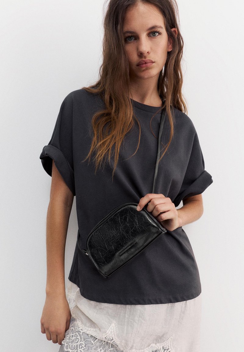Jeune femme aux longs cheveux porte une chemise sombre à manches retroussées et tient un petit sac bandoulière en cuir noir au-dessus d'une jupe blanche en dentelle.