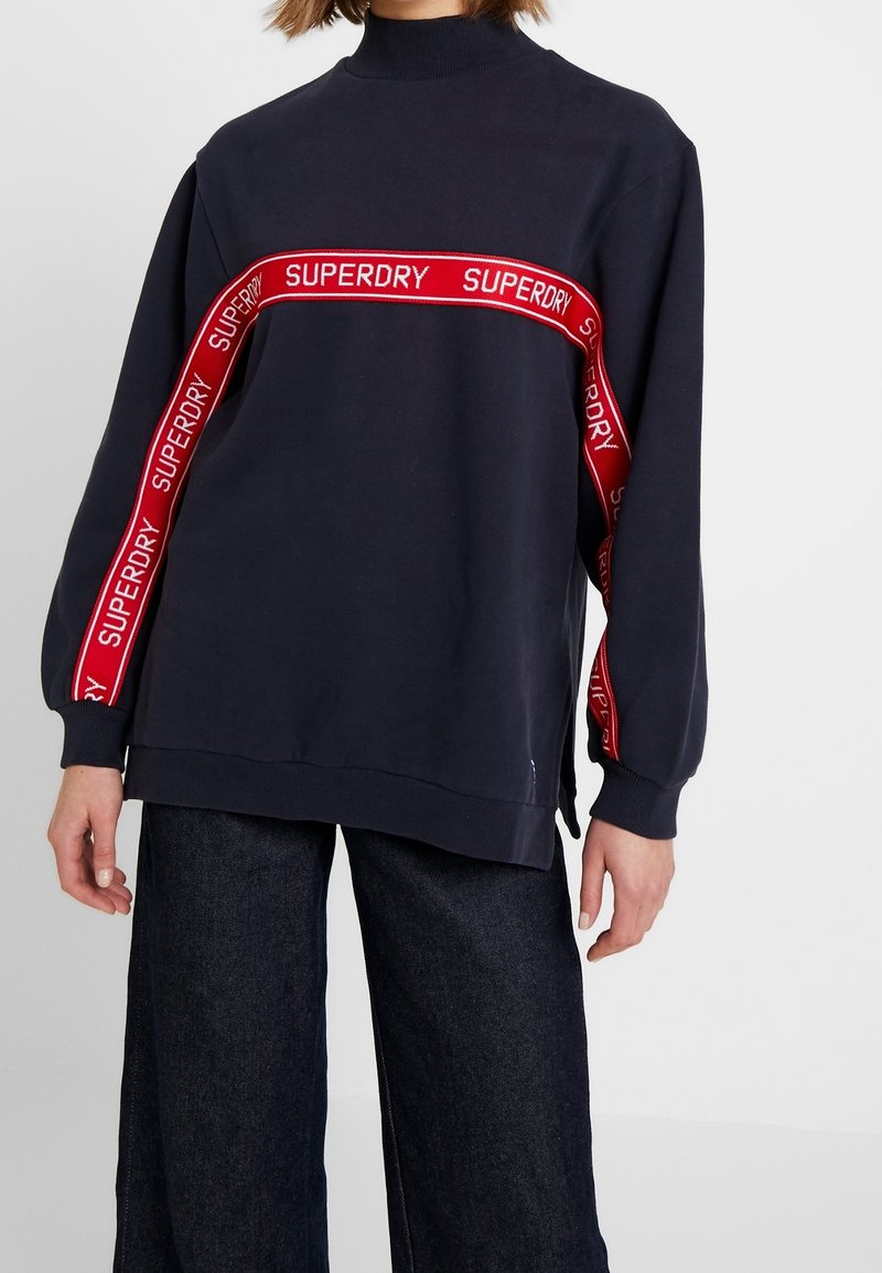 Personne portant un sweat-shirt bleu marine avec une bande diagonale rouge et le texte blanc « SUPERDRY » sur la poitrine et les manches, associé à un pantalon foncé.
