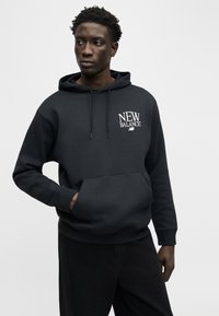 Νεαρός άνδρας που φοράει μαύρο φούτερ New Balance με μπροστινή τσέπη, κοιτάζοντας προς τα αριστερά του, σε απλό ανοιχτό φόντο.