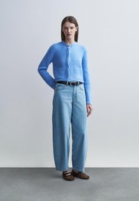 Donna in piedi che indossa un cardigan azzurro peloso, jeans a gamba larga a vita alta di colore azzurro chiaro con texture, cintura nera e scarpe basse con stampa leopardata.