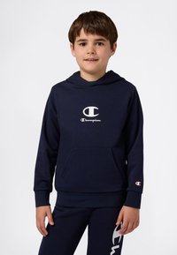 Ragazzino che indossa un hoodie Champion blu navy con tasca frontale e pantaloni abbinati con logo sulla coscia, in piedi davanti a uno sfondo bianco.