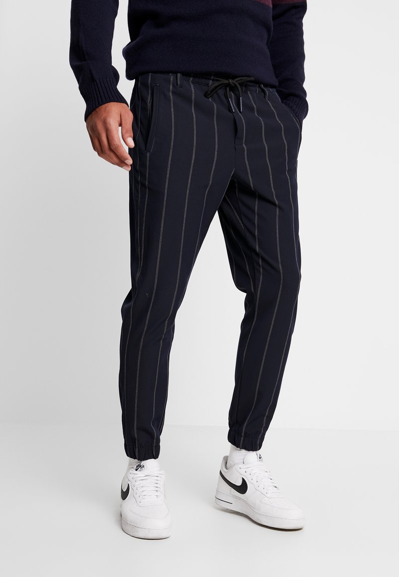 Homme portant un pantalon de jogging bleu marine avec de légères rayures verticales, des baskets blanches Nike et un pull sombre, se tenant devant un fond uni clair.